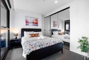 A modern hotel room with a large bed, a nightstand with a lamp, a large mirror, and artwork on the walls.