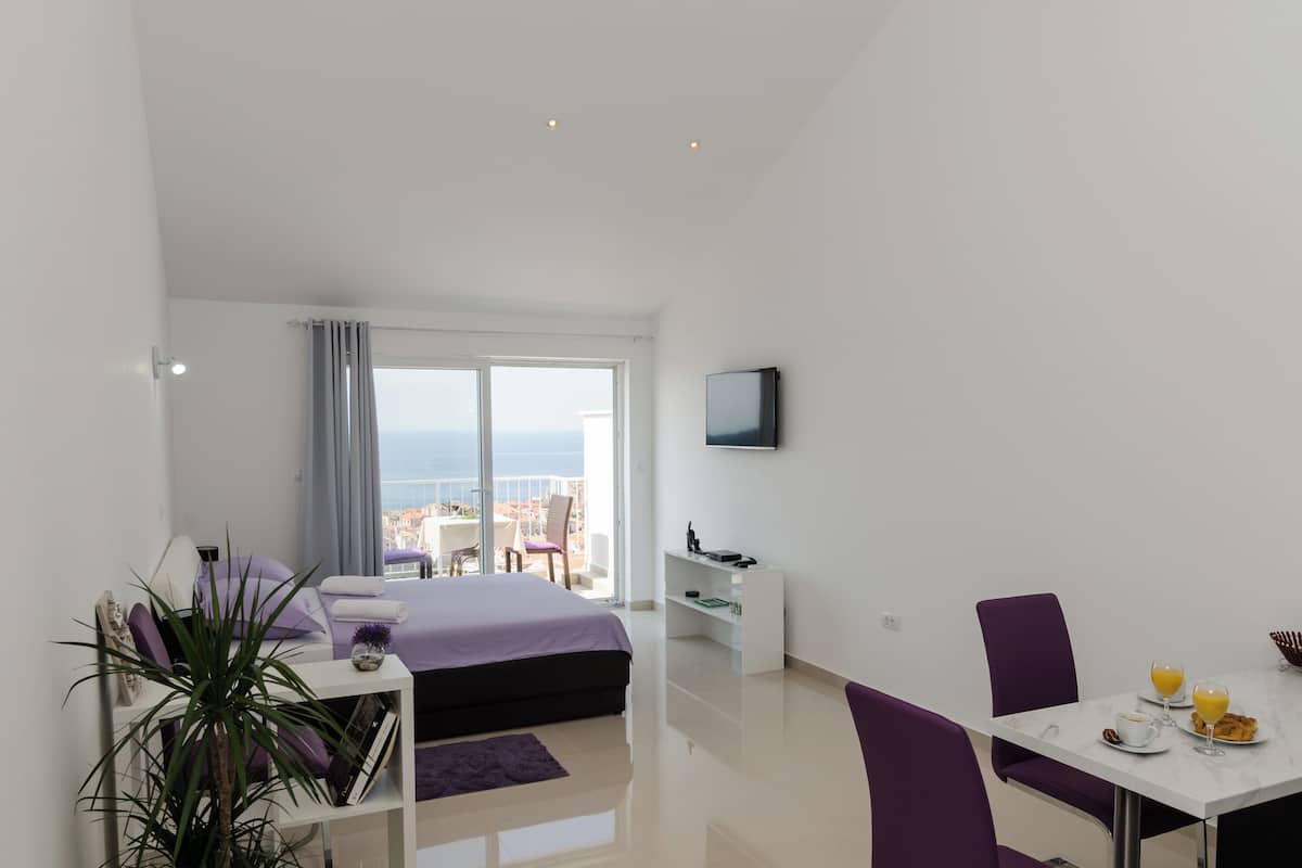 A modern hotel room with a balcony, a bed, a desk, and a dining area with breakfast.