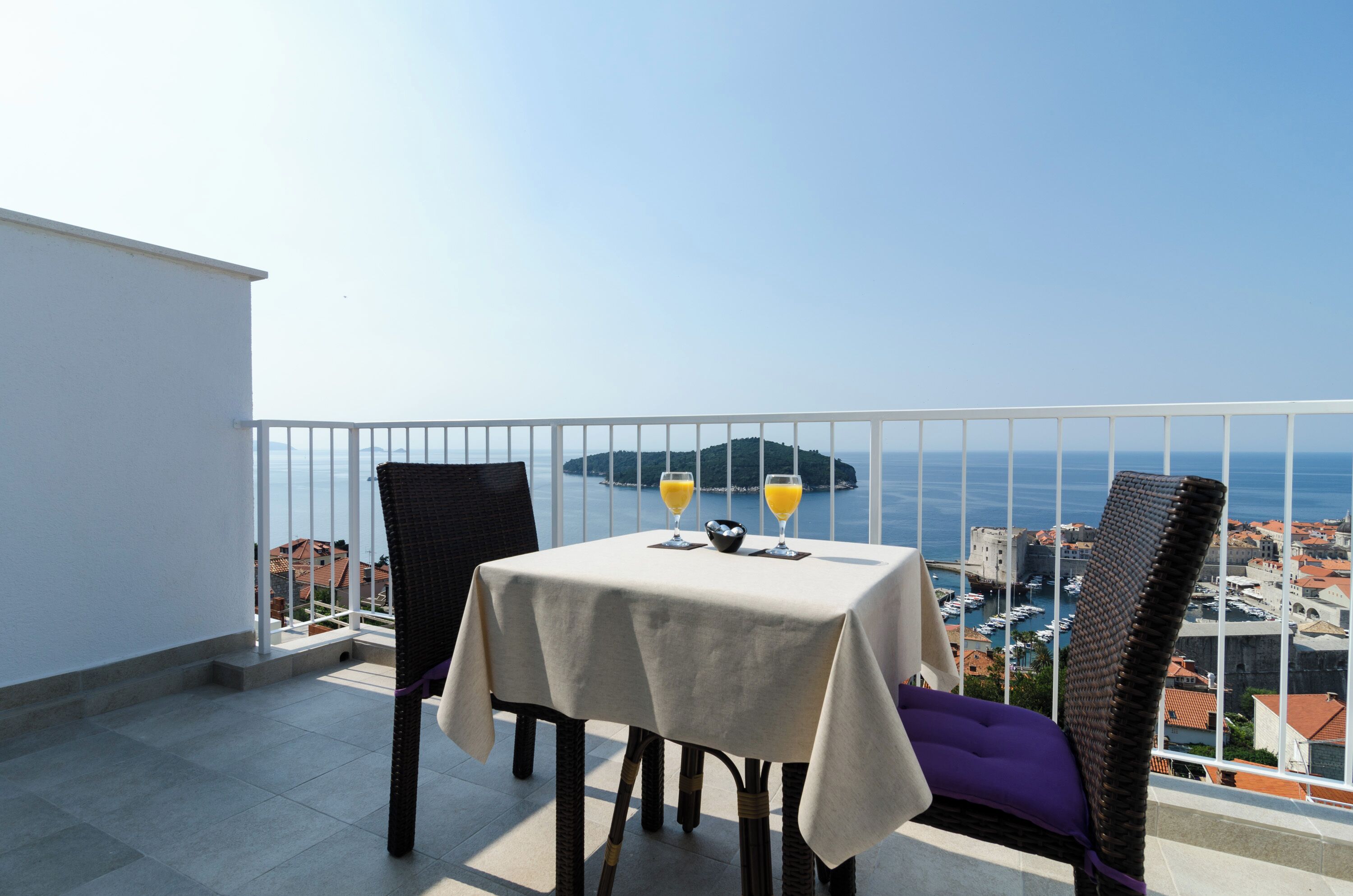 A balcony with a table and chairs, two glasses of orange juice, and a view of the sea and buildings.