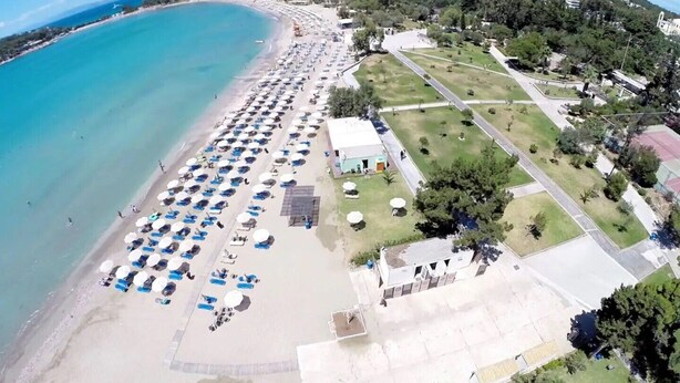 Ein Luftbild eines Strandes mit Reihen von Sonnenschirmen und Liegestühlen, einem Parkplatz und einer Grünfläche mit Wegen.