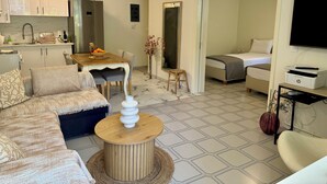 Ein modernes Wohnzimmer mit Sofa, Couchtisch und Essbereich. Es gibt eine Küche mit Schränken und einem Kühlschrank. Im Hintergrund ist ein Bett zu sehen.