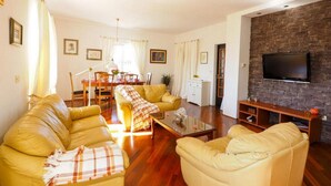 Una sala de estar con dos sofás amarillos, una mesa de centro, un televisor montado en una pared de piedra y un comedor con mesa y sillas.
