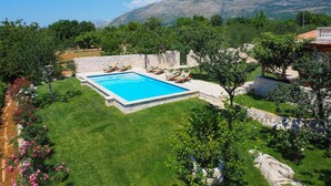 Una piscina rectangular con sillas de descanso, rodeada de césped cuidado y muros de piedra, con montañas al fondo.