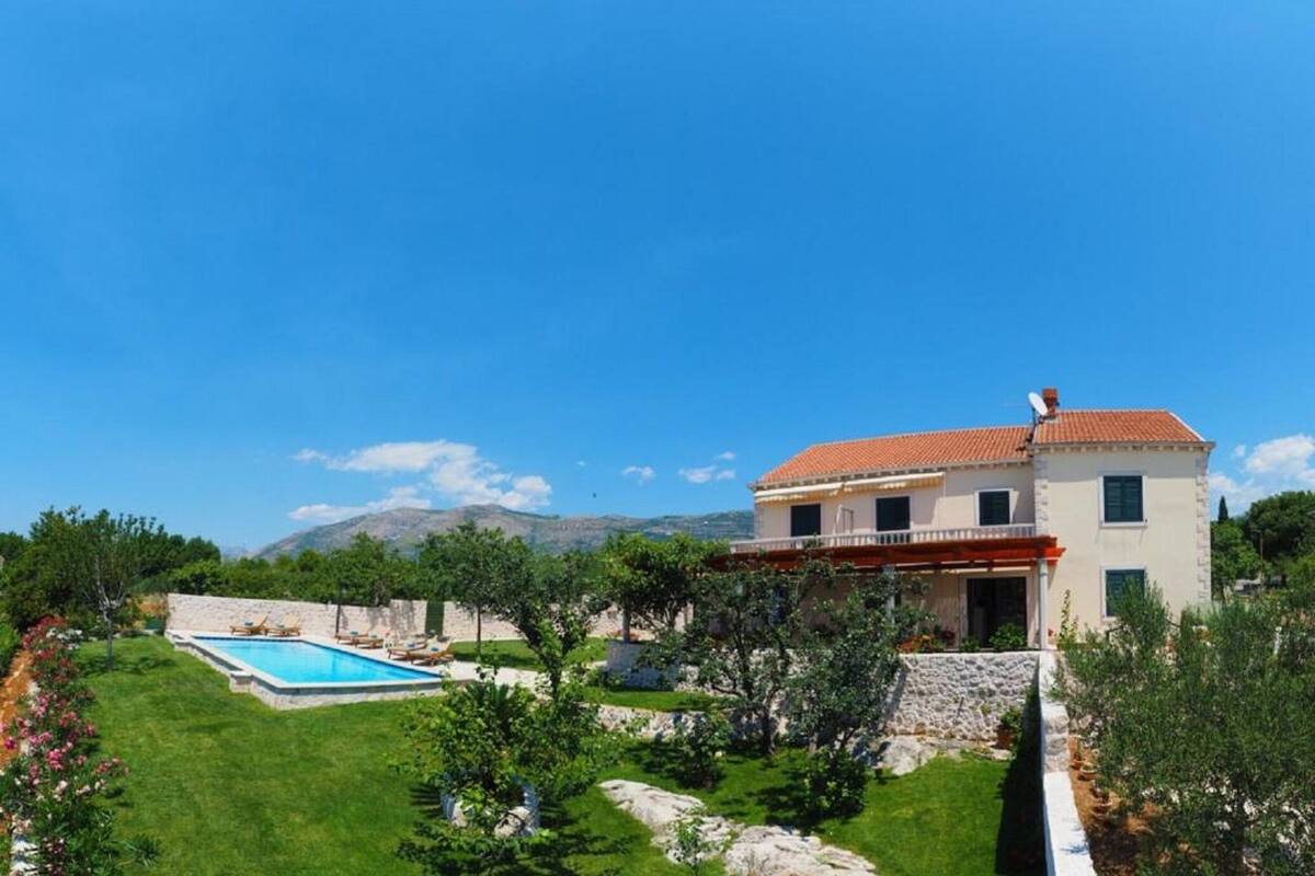 A two-story house with a red-tiled roof, a swimming pool, and a stone wall.
