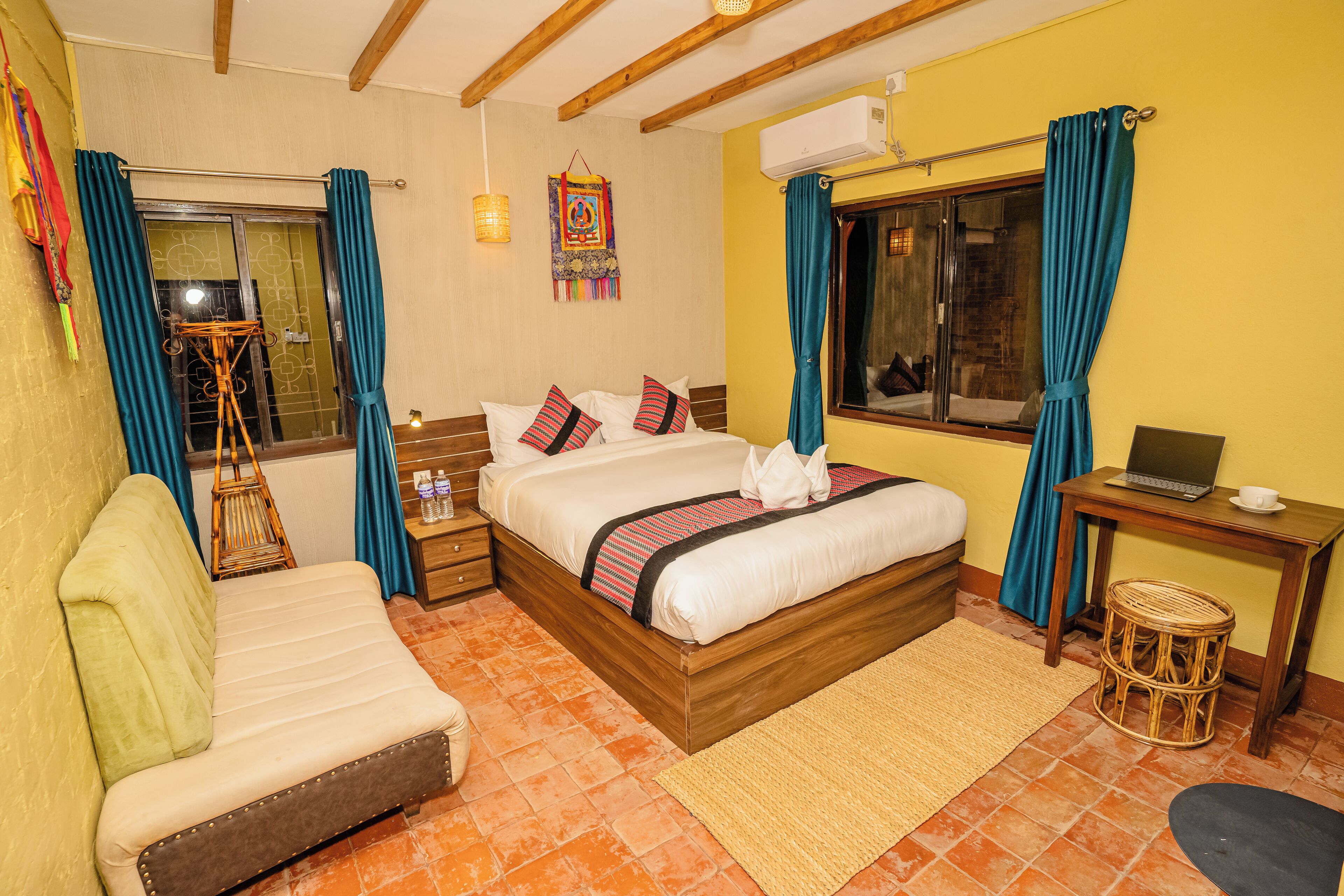Habitación de hotel con cama, sofá, una pequeña mesa con una computadora portátil, una cesta de mimbre y una ventana con cortinas.