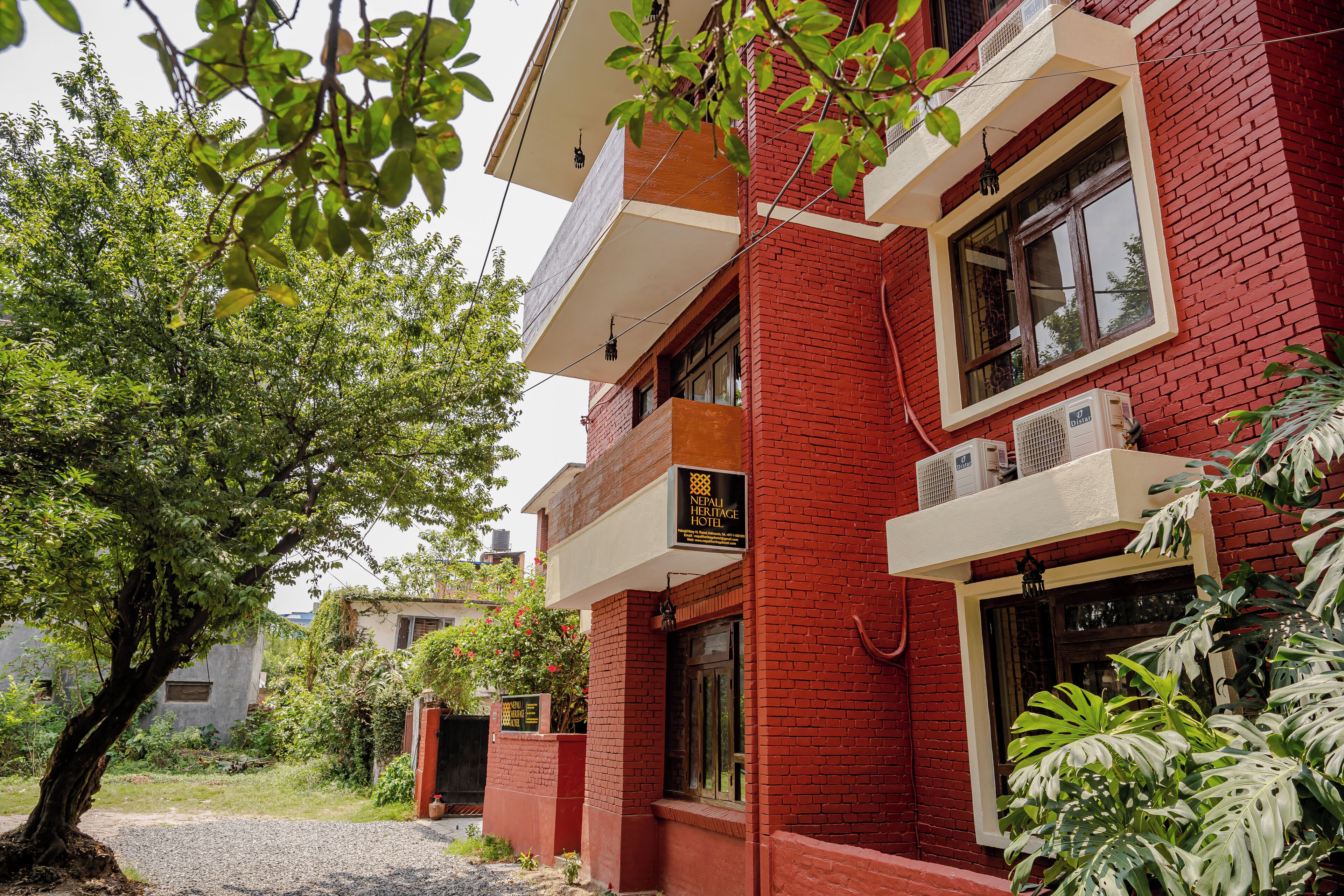 A red brick building with a sign for the Nepal Heritage Hotel. There are trees and a gravel path in front.