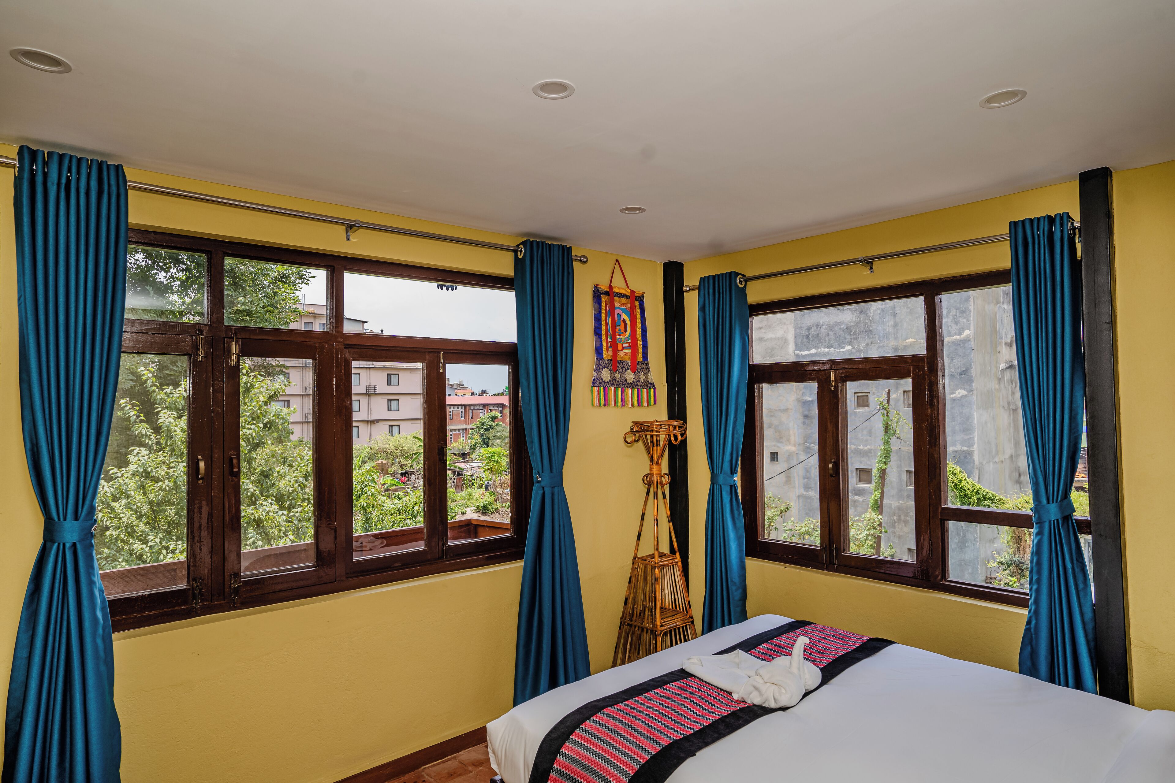 A room with a bed, two windows with curtains, a wall-mounted decoration, and a view of buildings outside.