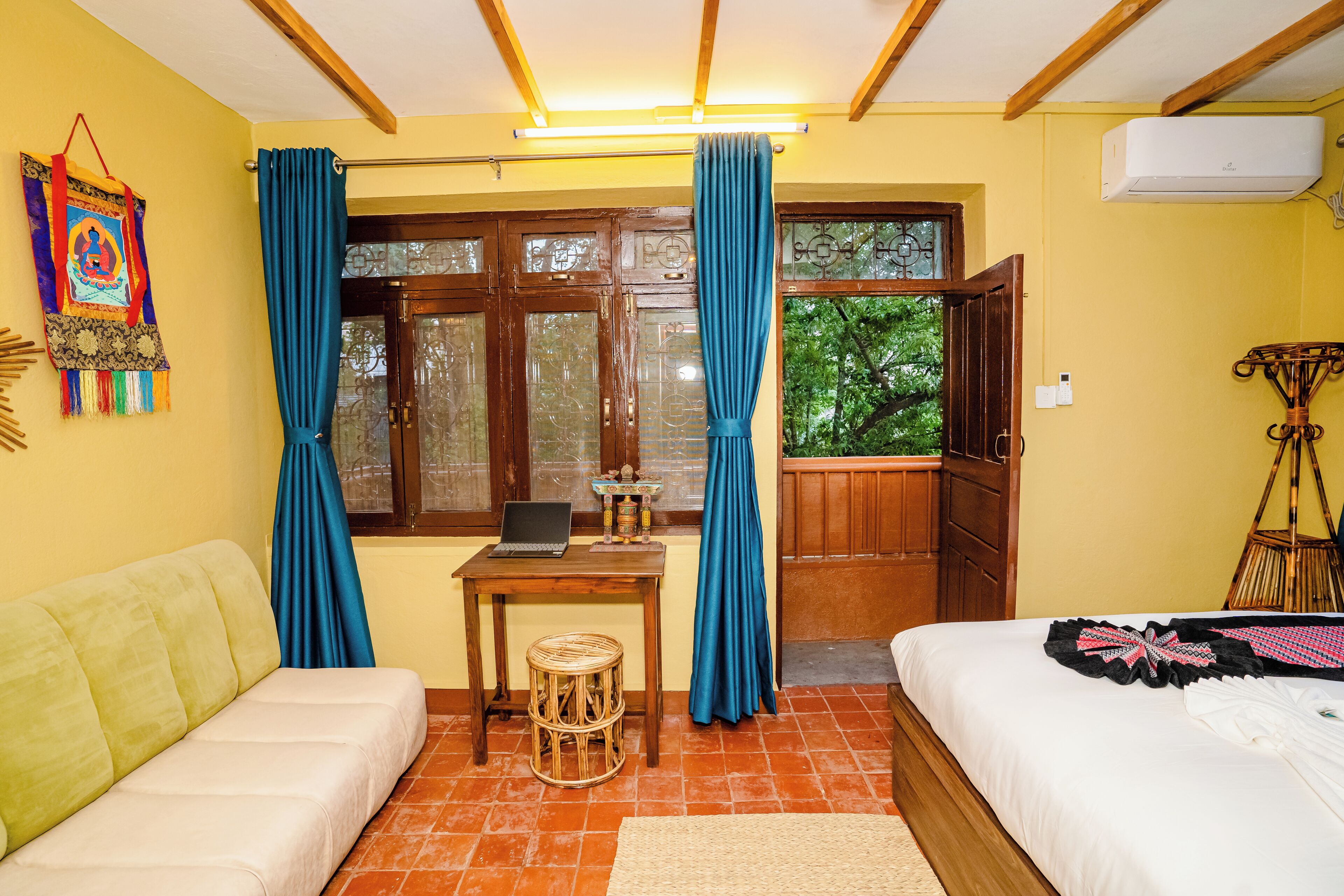 A room with a bed, a sofa, a small table with a laptop, a window with curtains, and an air conditioning unit.