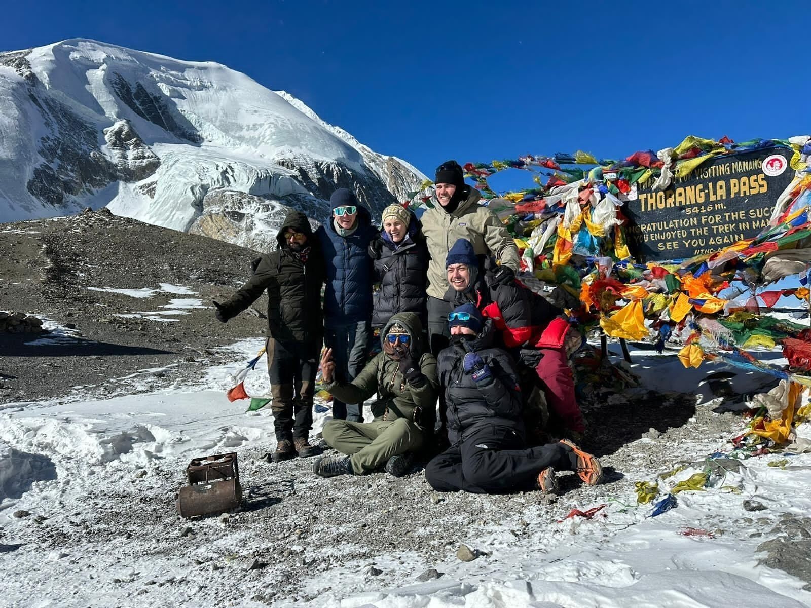 A group of people posing in front of a sign with colorful prayer flags, with a snow-covered mountain in the background.