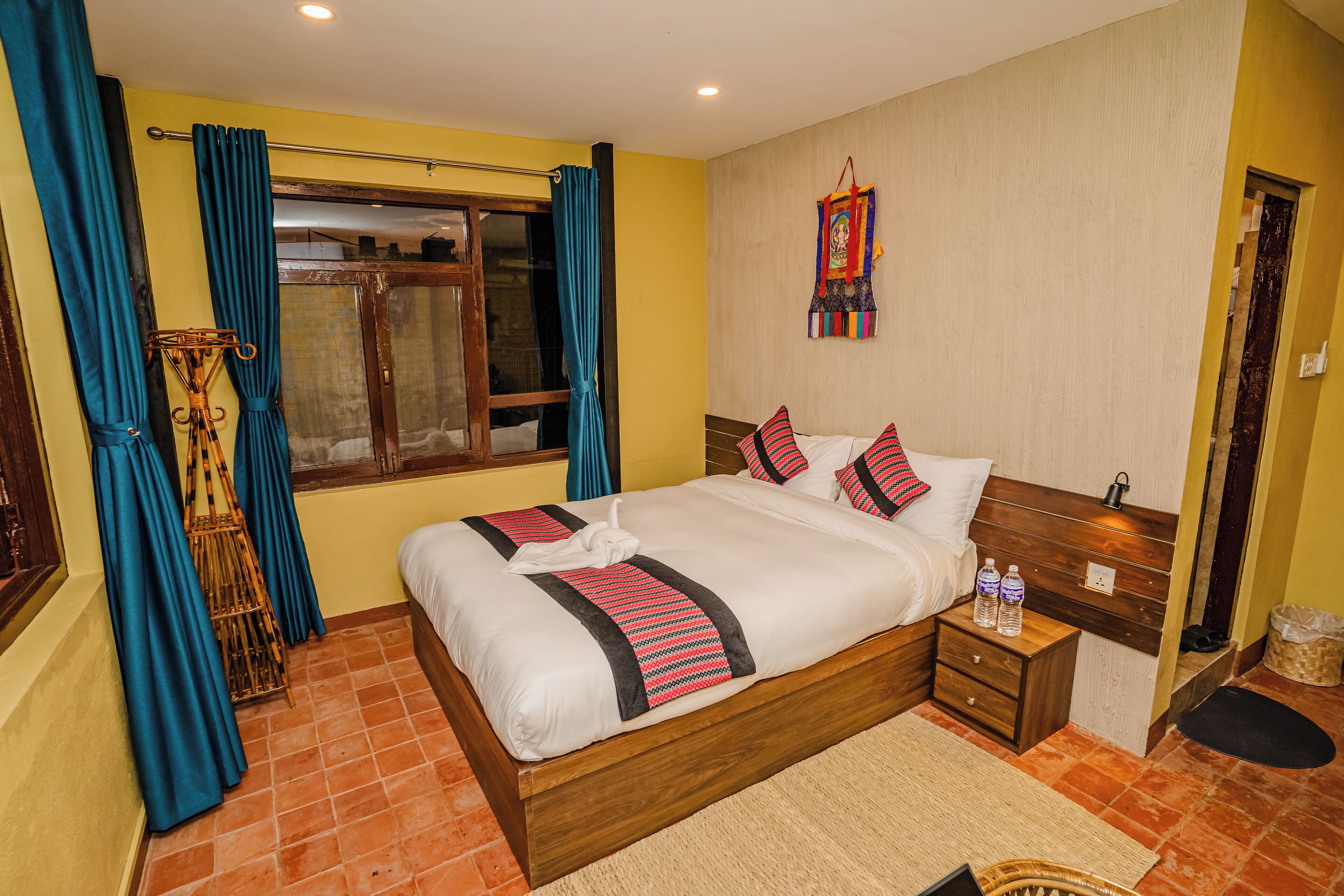 A neatly arranged hotel room with a wooden bed, a bedside table with water bottles, a small rug, and a window with curtains.