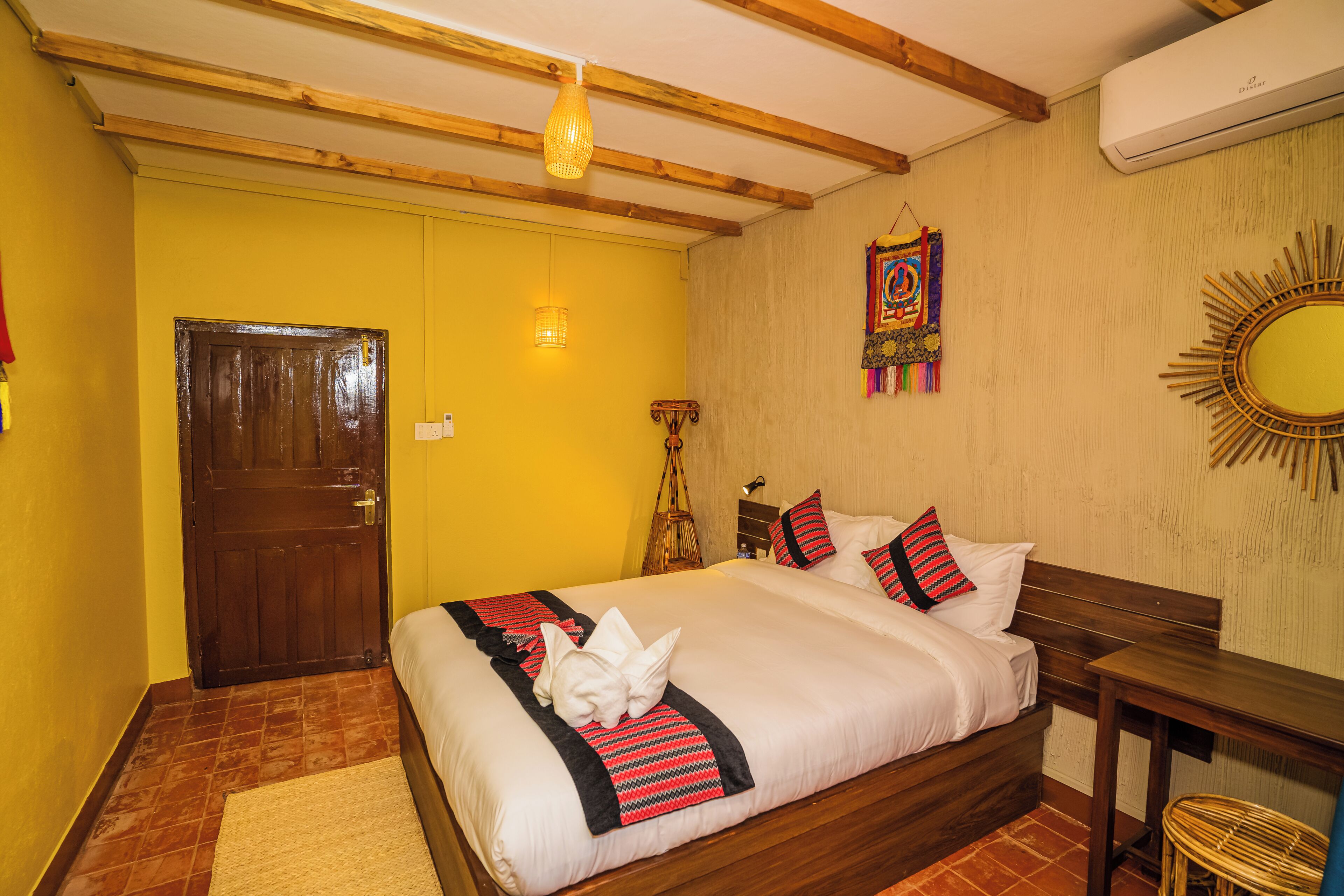 A hotel room with a wooden bed, a small table, a wall-mounted mirror, and a colorful wall hanging.