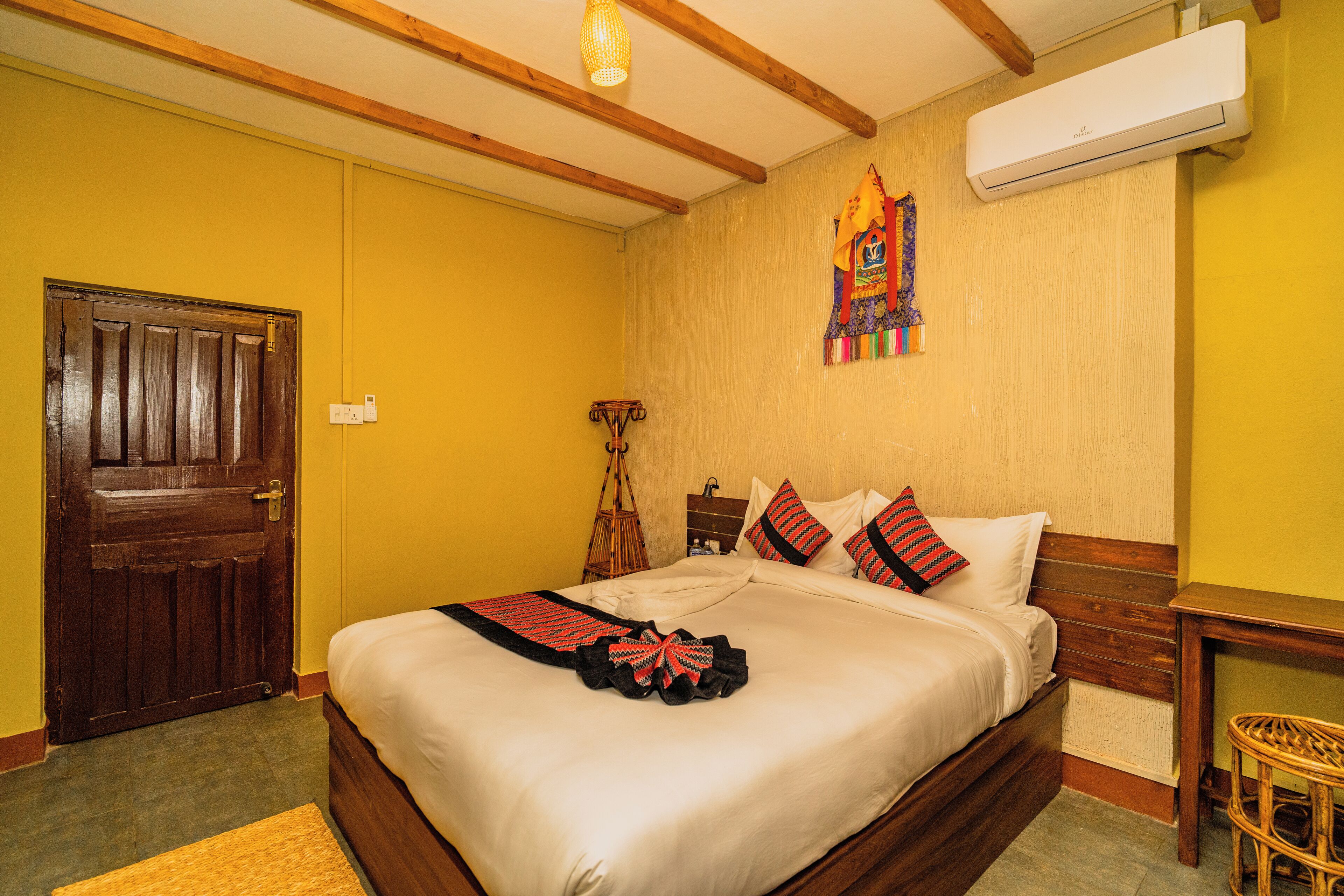 Una habitación de hotel con estructura de cama de madera, una cama grande con ropa de cama blanca, cabecera de madera, aire acondicionado montado en la pared, una puerta de madera y una pequeña silla de mimbre.