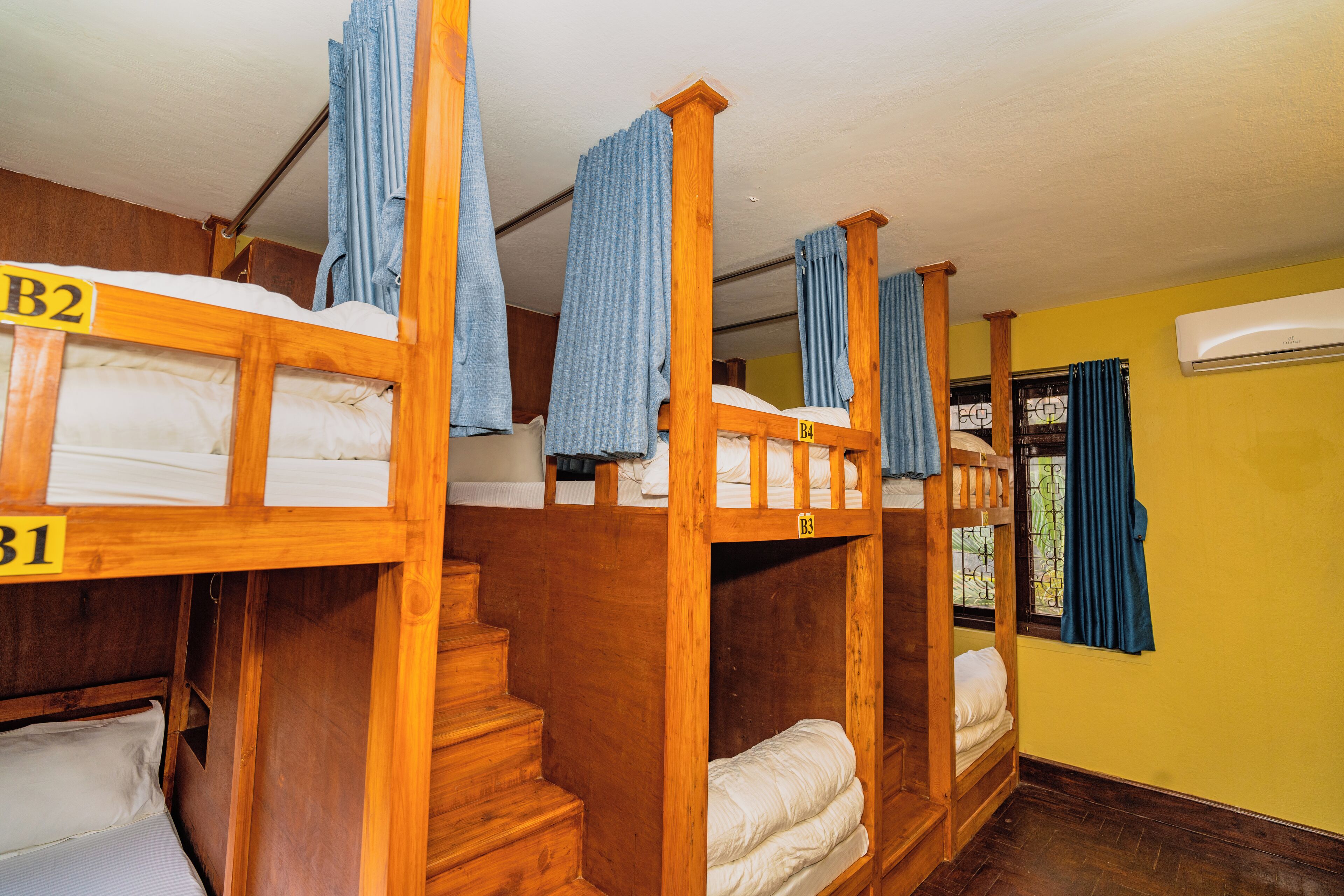 A dormitory room with bunk beds labeled B1, B2, and B3, wooden stairs, and a window with curtains.