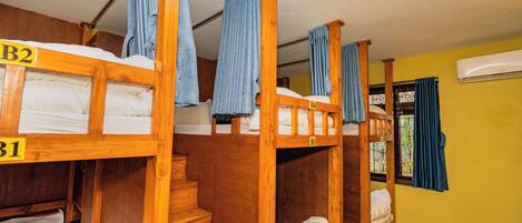 A dormitory room with bunk beds labeled B1, B2, and B3, wooden stairs, and a window with curtains.