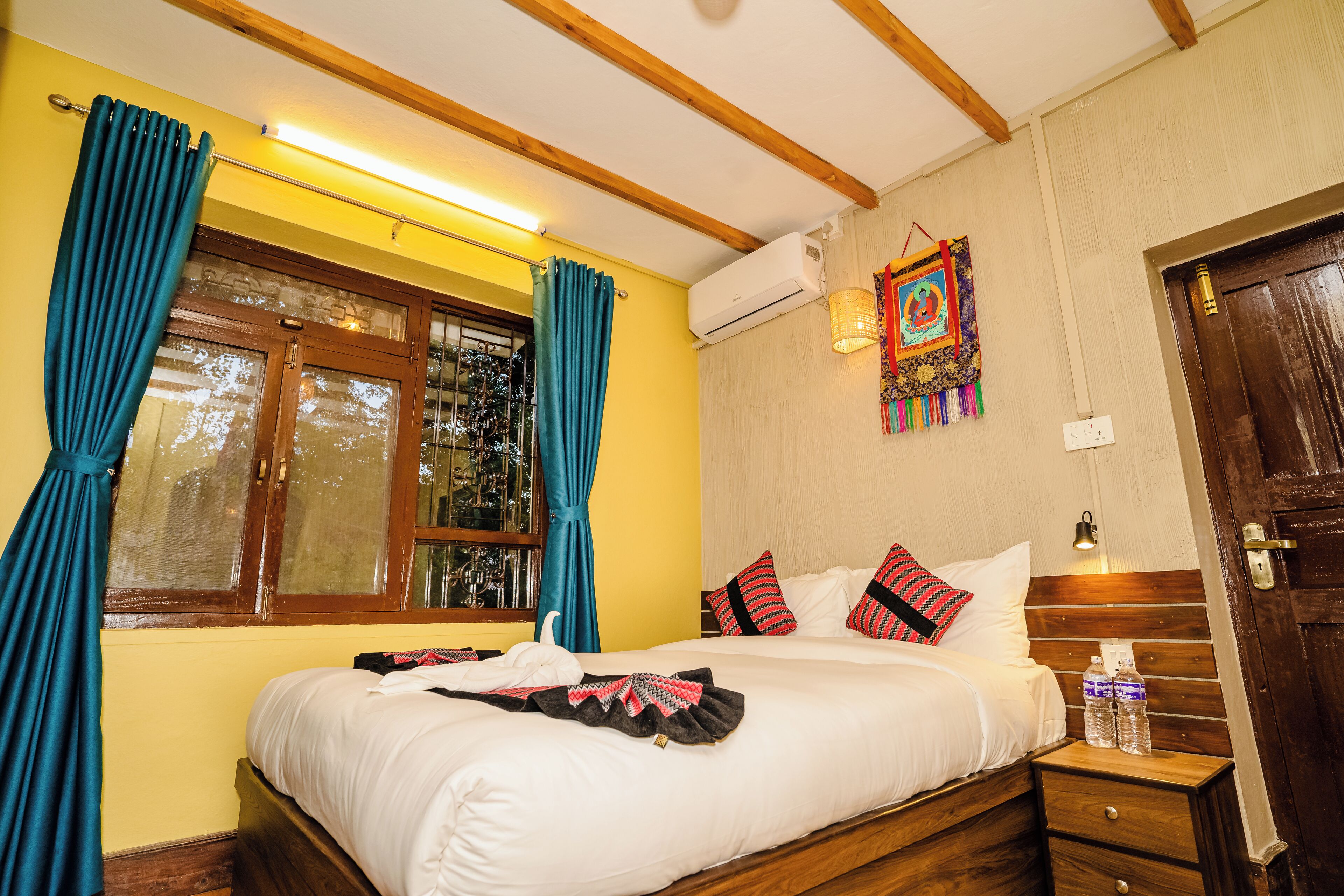 Habitación de hotel con una cama grande, cabecero de madera, una ventana con cortinas, aire acondicionado de pared, una lámpara de noche y una mesita de noche de madera con una botella.