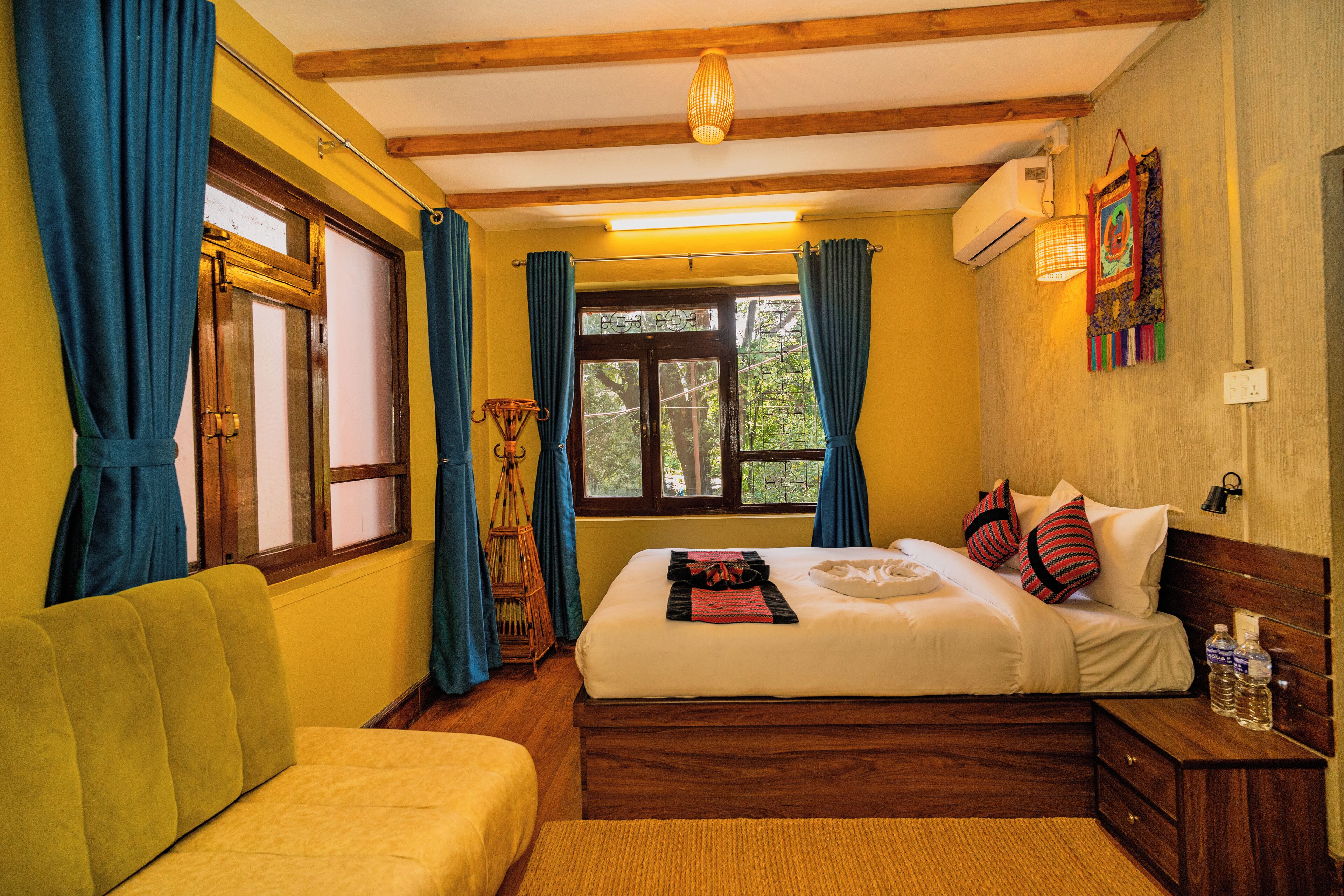 A room with a bed, a wooden headboard, a window with curtains, a wall-mounted air conditioner, and a small table with a lamp.