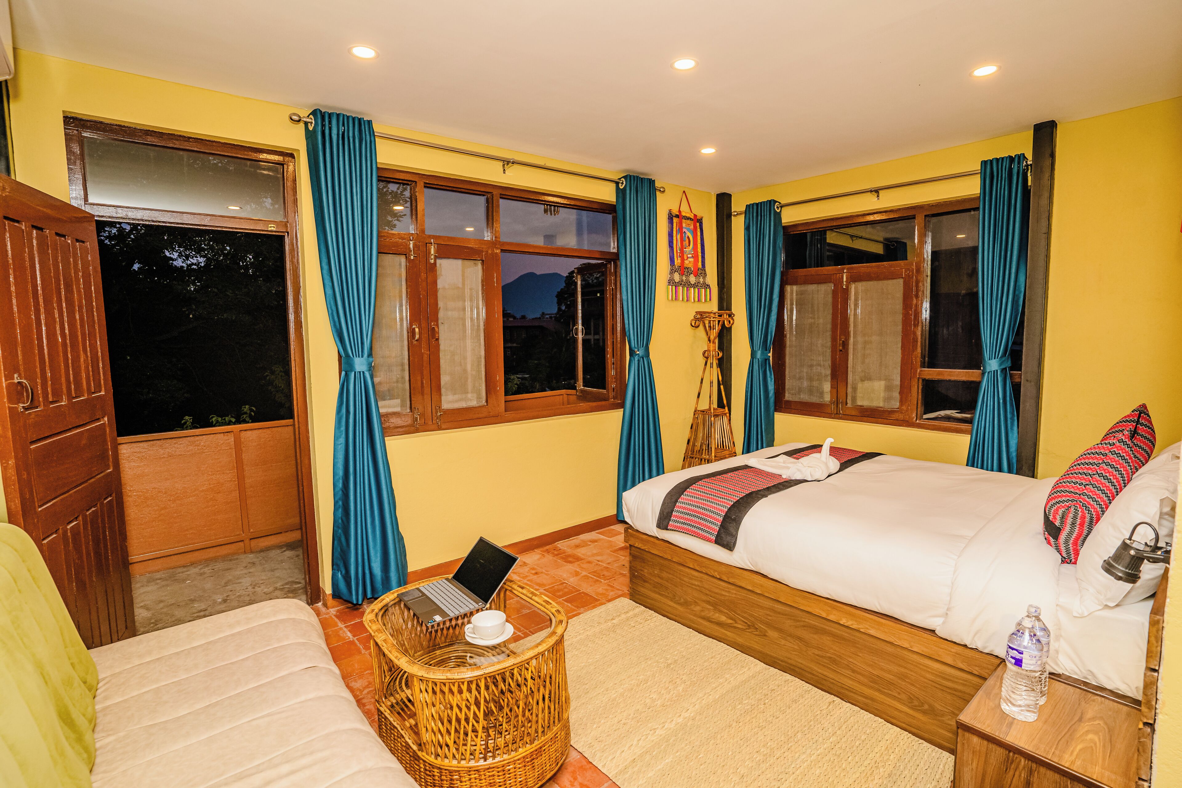 Una habitación con cama, una silla de mimbre, una mesita de noche y un ventanal con cortinas.