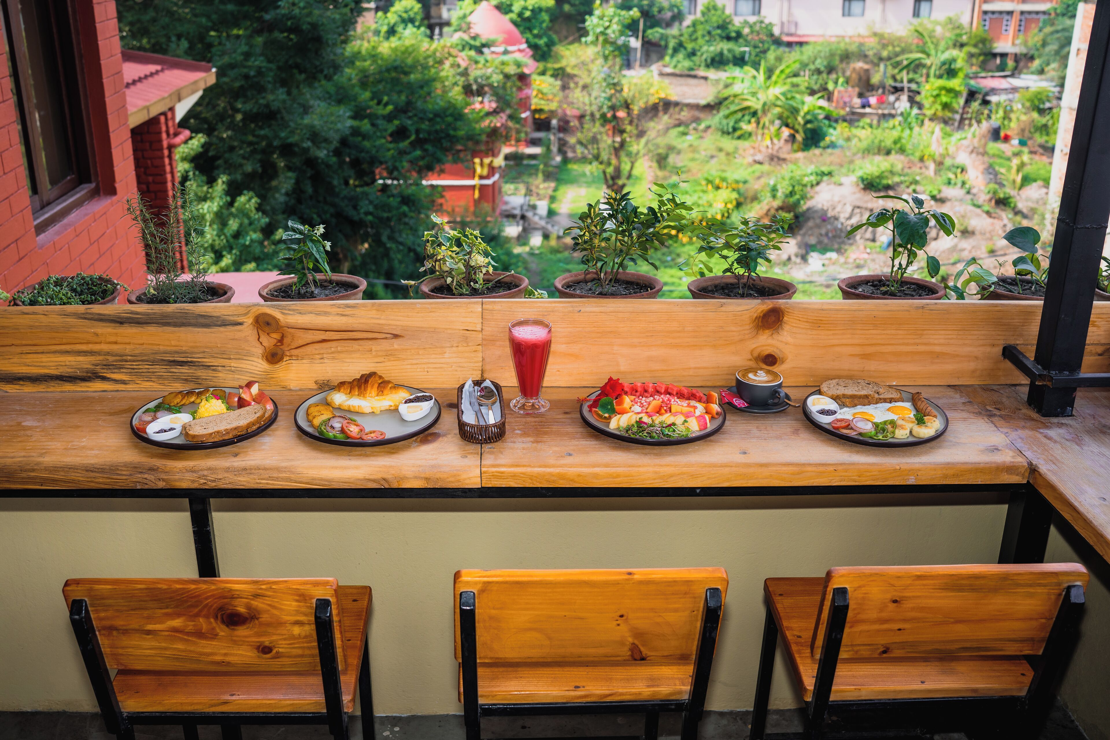 A wooden table with breakfast plates, a glass of red juice, and potted plants on a balcony overlooking a garden.