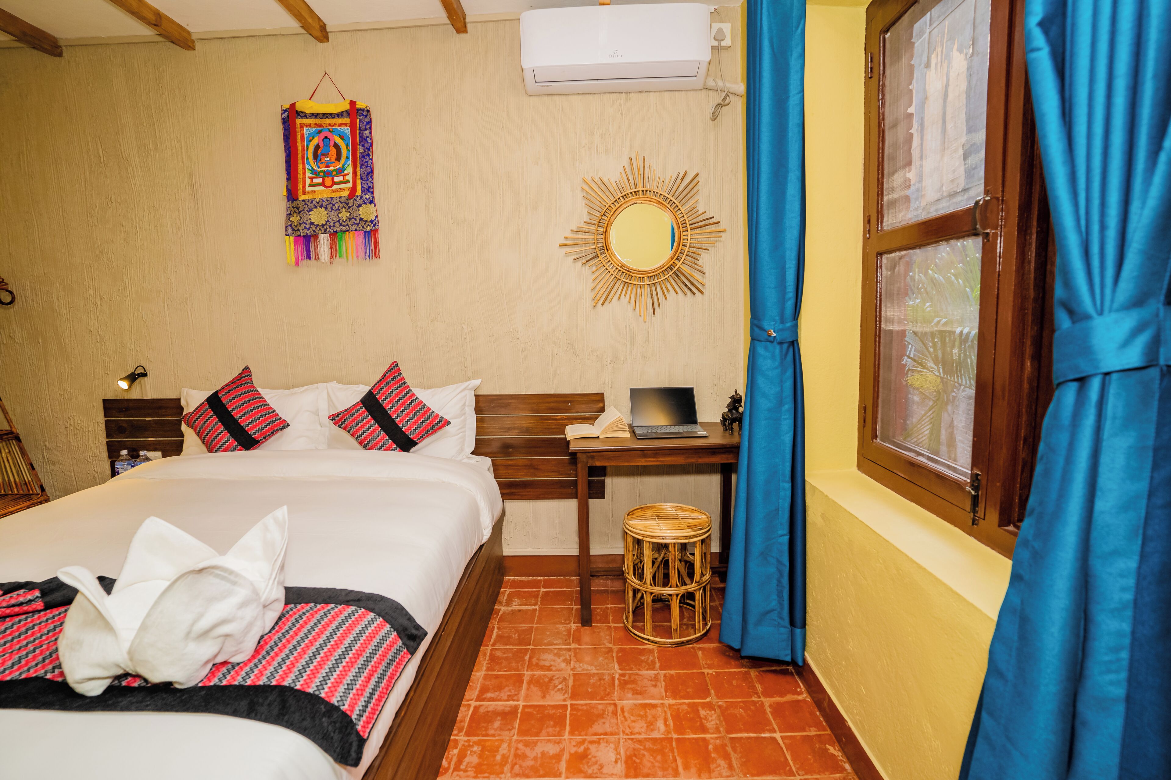 A neatly made bed with pillows, a small round table with a laptop, a wall-mounted mirror, and a window with curtains.