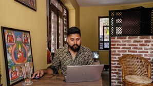 Una persona usando un ordenador portátil en una mesa de madera, con un vaso de vidrio transparente y un cuadro enmarcado en la pared.