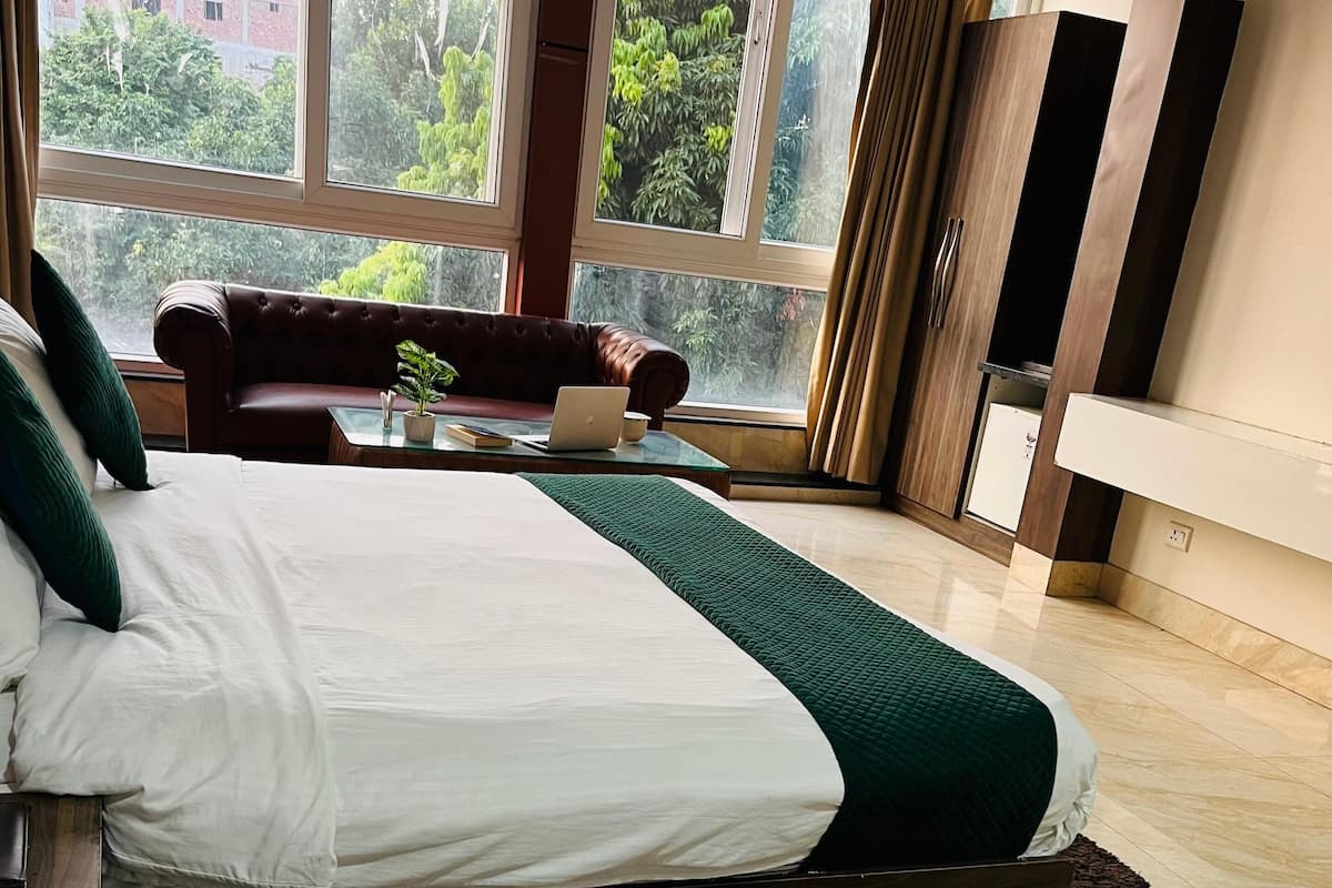 Une chambre d’hôtel moderne avec deux lits, un canapé, un ordinateur portable posé sur une table en verre, et de grandes fenêtres offrant une vue sur la verdure et les bâtiments.