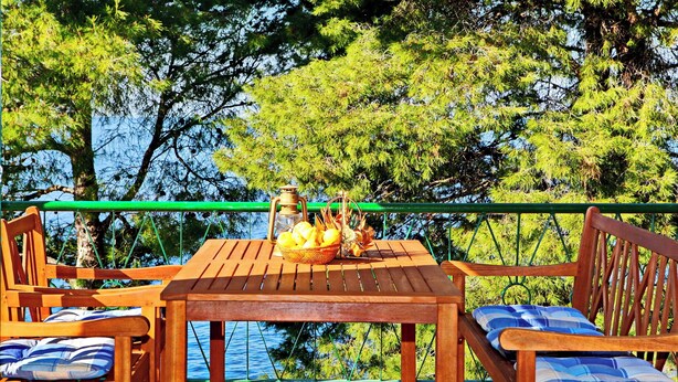 Un tavolo e delle sedie in legno con un cesto di frutta e una bottiglia, affacciati su uno specchio d'acqua con alberi sullo sfondo.