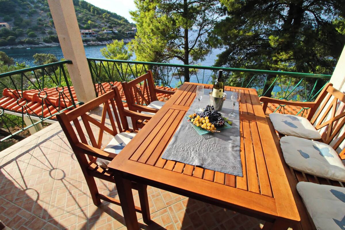 Una mesa de madera dispuesta con una botella, vasos y un bol de fruta en una terraza con vista a un lago y árboles.