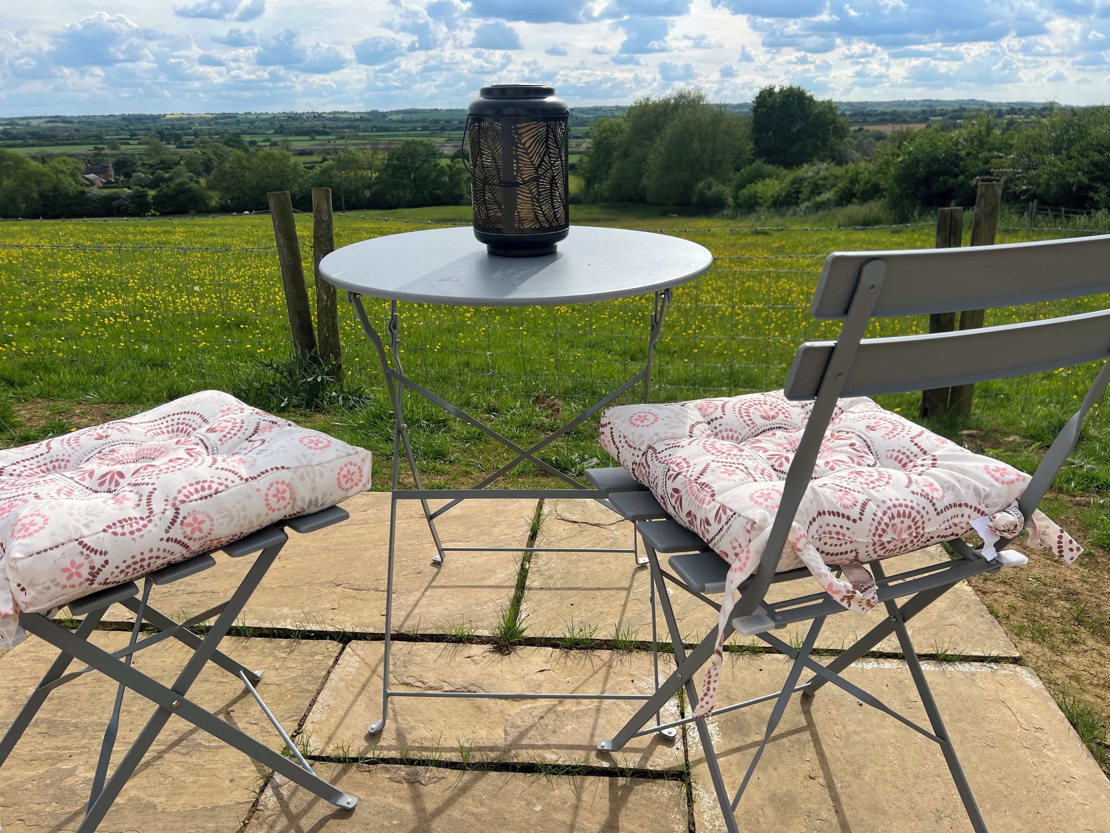 An outdoor seating area with a table and two chairs, a decorative lantern, and a view of a green field and distant landscape.