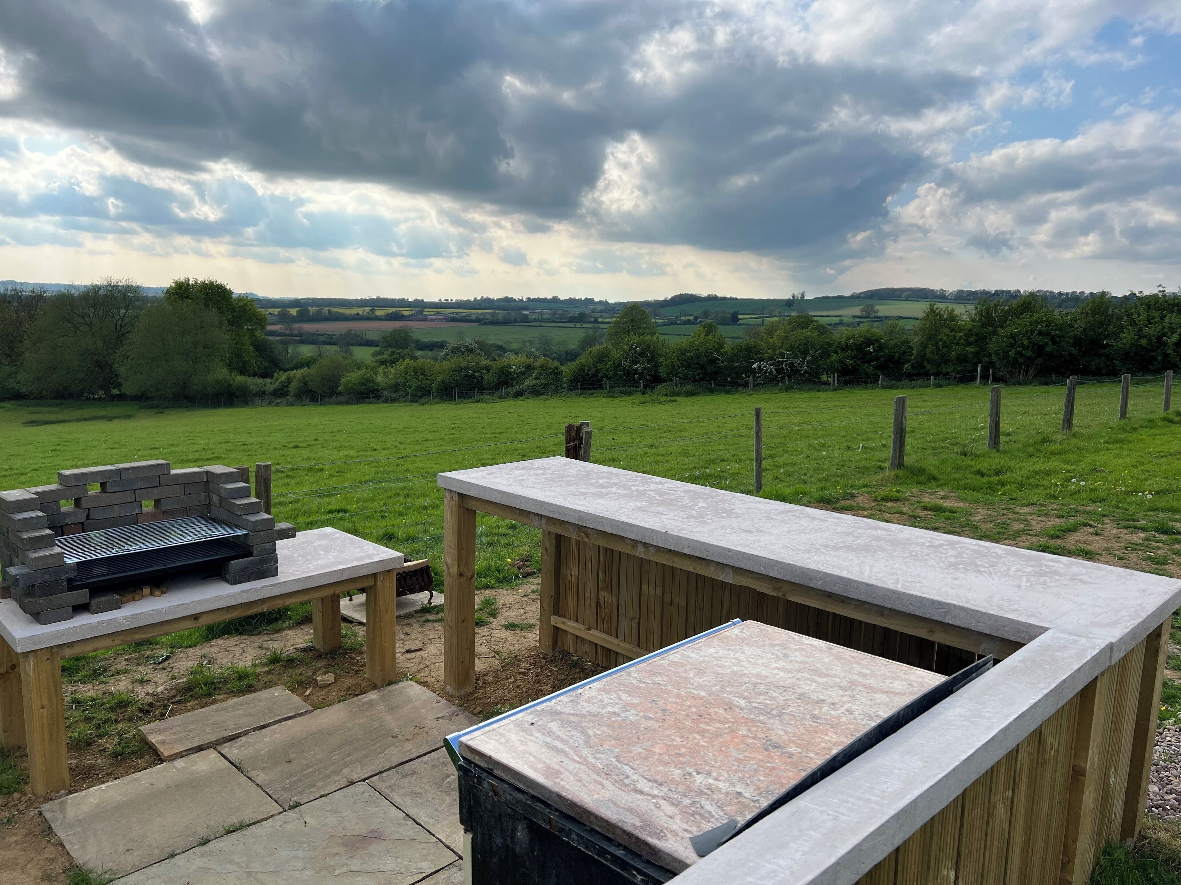 Un espace extérieur aménagé en terrasse, avec une table en pierre et un barbecue, entouré d’une clôture en bois et offrant une vue sur un champ verdoyant.