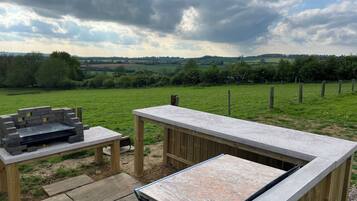 Un espace extérieur aménagé en terrasse, avec une table en pierre et un barbecue, entouré d’une clôture en bois et offrant une vue sur un champ verdoyant.