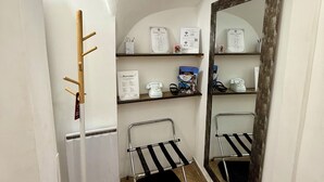 Un pasillo estrecho con un perchero, una silla pequeña y un espejo en la pared. Estantes arriba albergan varios objetos, incluyendo un teléfono, una caja y algunos papeles.