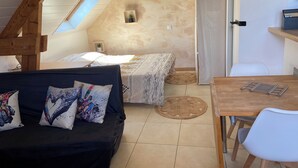 A cozy room with a sloped ceiling, a bed with patterned bedding, a black sofa with colorful pillows, a wooden table, and a small potted plant. - Studio à la Campagne. Situé à 25 Minutes des Plages de Pénestin, Damgan (Missillac)