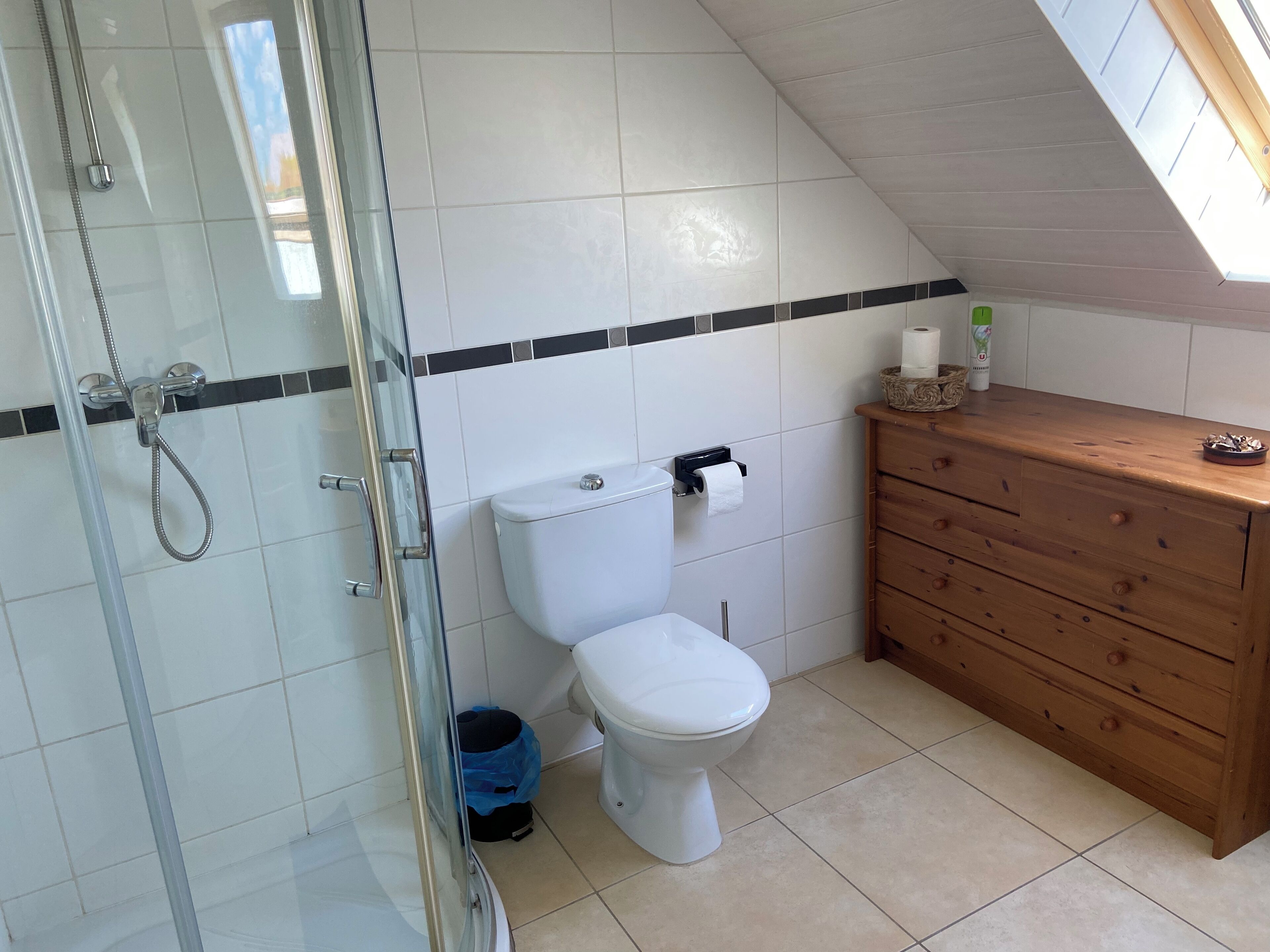 Een kleine, moderne badkamer met een douchecabine van glas, een wit toilet en een houten wastafelmeubel met een mand vol toiletartikelen.