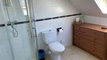 Een kleine, moderne badkamer met een douchecabine van glas, een wit toilet en een houten wastafelmeubel met een mand vol toiletartikelen.
