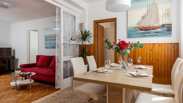 A dining area with a wooden table, white chairs, and a red sofa. There is a painting of a sailboat on the wall and a potted plant on the table. - Garden Vista Apartment (Dubrovnik)
