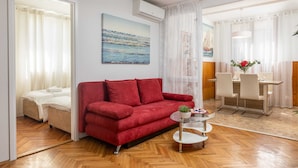 A living room with a red sofa, a small round table, and a painting on the wall. - Garden Vista Apartment (Dubrovnik)