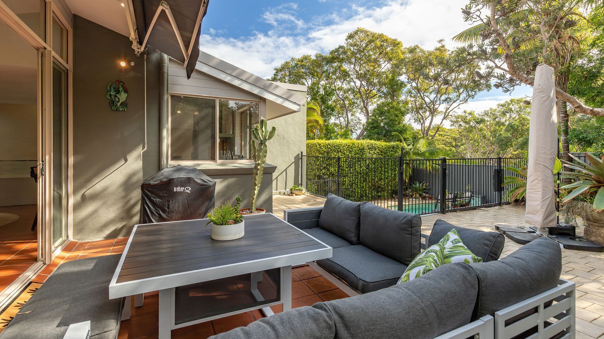 A patio with a table and sofa, a barbecue grill, and a pool in the background.