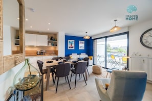 Uma cozinha moderna com armários brancos, área de jantar com mesa e cadeiras, parede azul de destaque e uma grande janela com vista para o exterior.