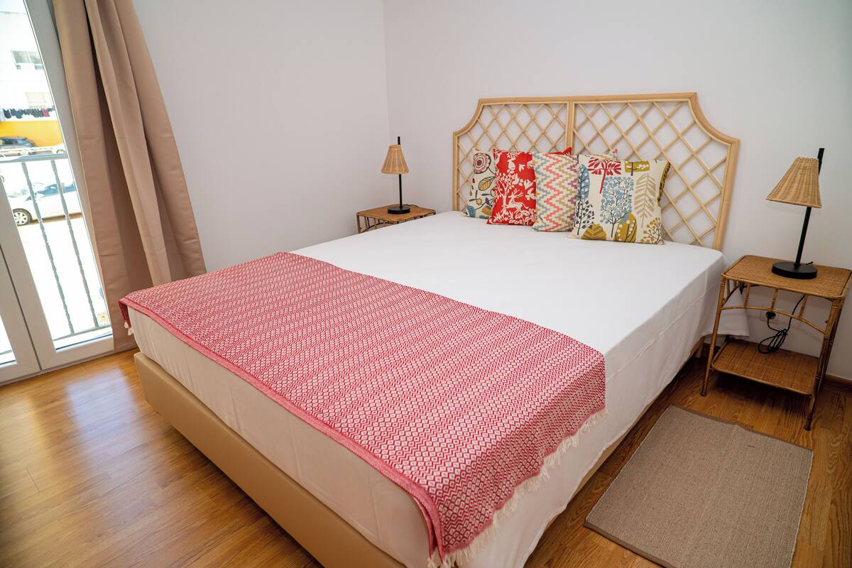 Una cama bien hecha con un edredón estampado, un cabecero de madera, dos mesitas de noche con lámparas y vistas al exterior a través de una ventana con cortinas.