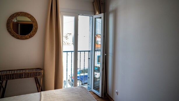 A bedroom with a bed, a wicker chair, a round mirror, and a sliding glass door leading to a balcony with a view of the outside. - Turisalentejo  (Redondo)
