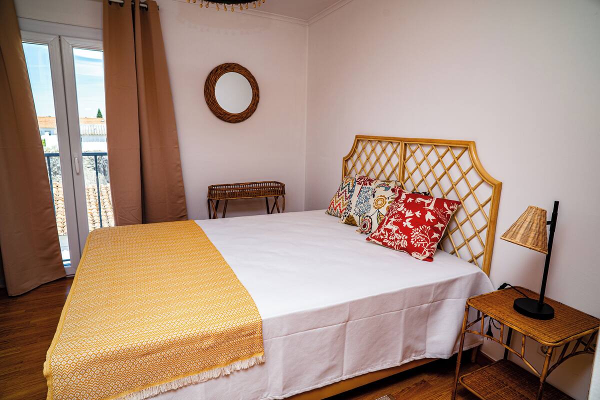 Una cama bien hecha con una colcha blanca y amarilla, un cabecero de madera y una mesita de noche con una lámpara.