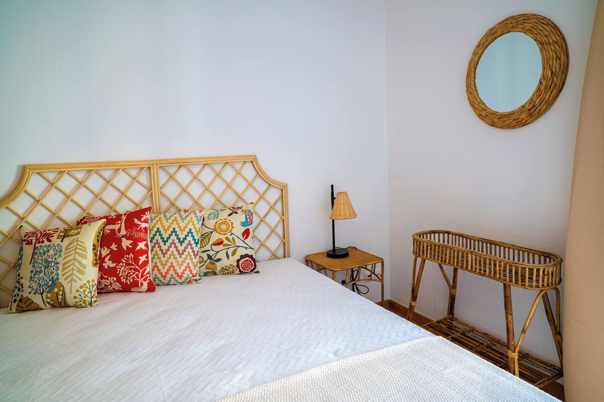 Una cama bien hecha con cabecera de madera, una mesita de noche con lámpara y una mesita auxiliar de mimbre.