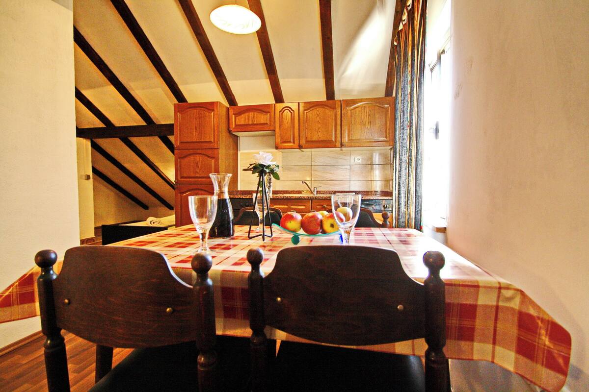A dining area with a table set for two, wooden chairs, a vase with flowers, and a fruit bowl.