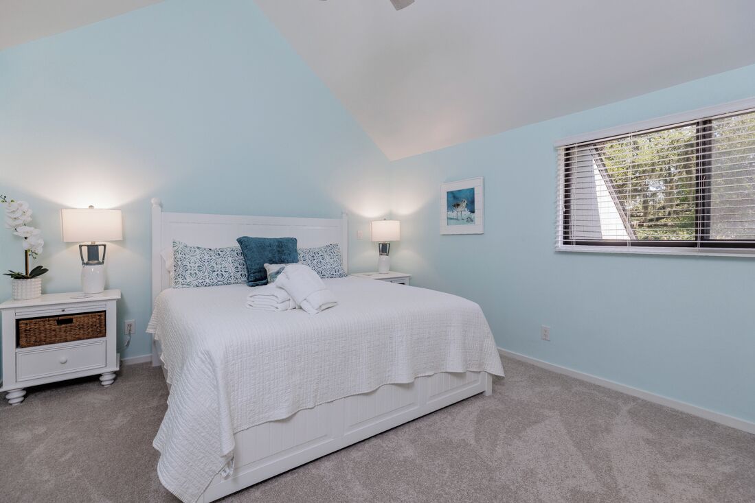 A bedroom with a white bed, two bedside tables, a window with blinds, and a wall-mounted picture.