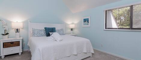 A bedroom with a white bed, two bedside tables, a window with blinds, and a wall-mounted picture.