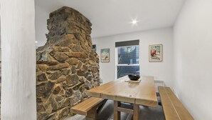 A stone fireplace, wooden dining table, and benches in a room with white walls and framed pictures.
