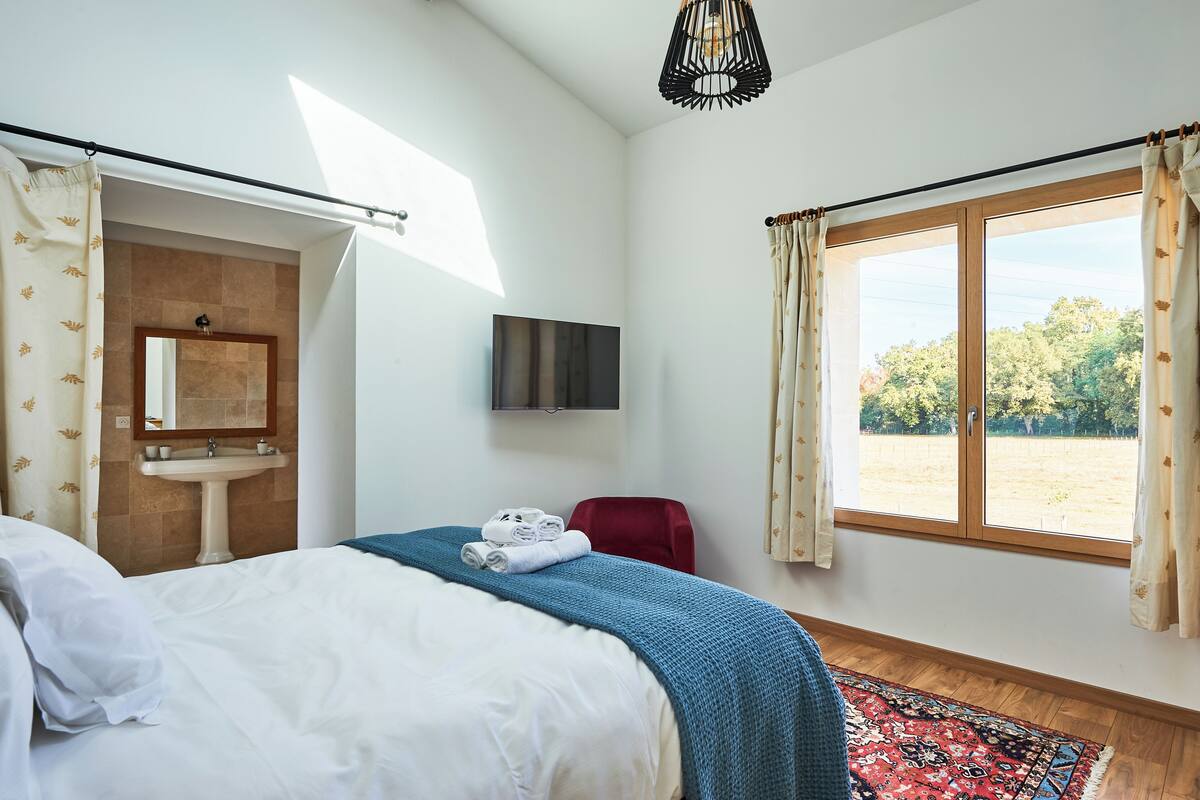 A bedroom with a bed, a red chair, a sink, a mirror, a TV, and a window with curtains.