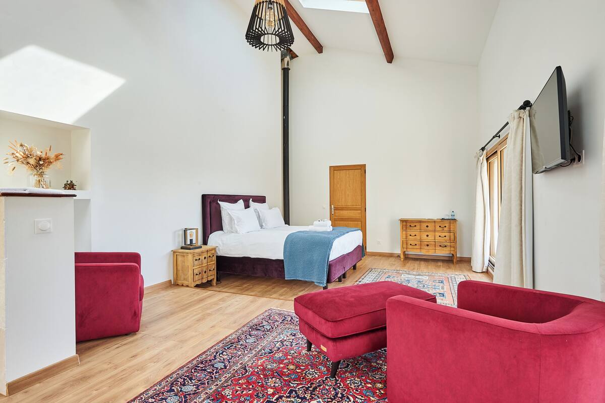 A bedroom with a bed, a red chair, a wooden dresser, a TV mounted on the wall, and a skylight.