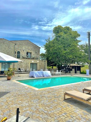 A pool area with stone flooring, a covered seating area, and a stone building in the background. - DOMAINE DE BADINE (Saint-Sulpice-et-Cameyrac)