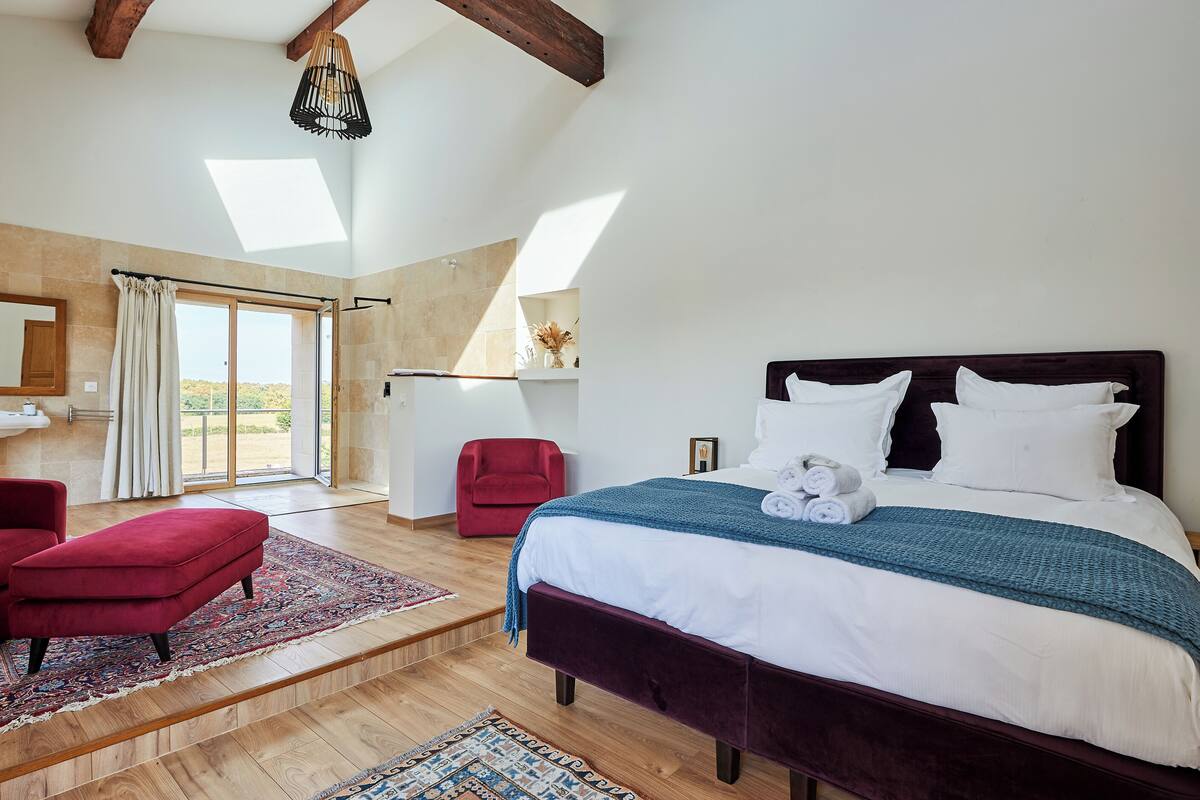 A bedroom with a large bed, a red chair, a small table, and a view of the outdoors.